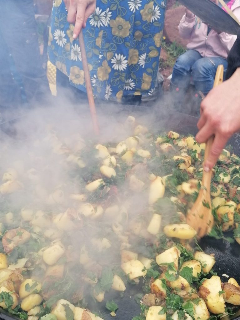 Eine große Pfanne, darin Kartoffeln und Wildkräuter, zwei Personen rühren in der Pfanne.